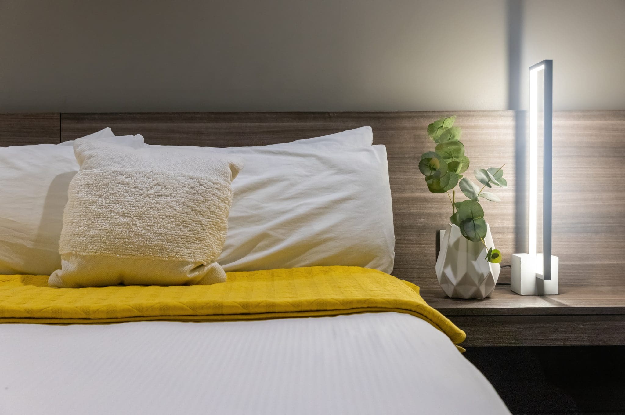 Close up shot of white pillows on a cozy, comfortable bed with an extra yellow blanket, with a plant and lamp on the side table in the Montvale Lofts at the Montvale Hotel in Spokane